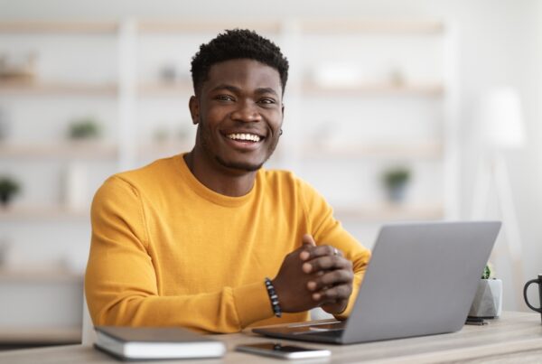 young man smiling looking at camera with hands folded in front of laptop